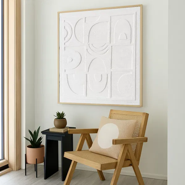 A cozy corner featuring a wooden chair and a small potted plant, with a textured white painting in a minimalist design hanging above.
