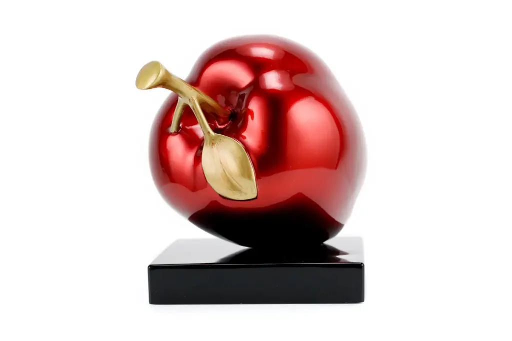 A sculptural piece of art depicting a glossy metallic red apple with a gold stem and leaf, resting on a black square base.